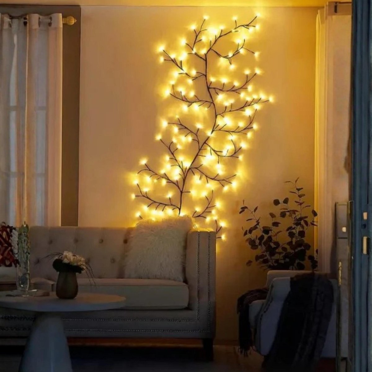 A decorative tree-shaped string light with warm yellow glowing bulbs mounted on a beige wall in a living room setting, with a sofa and table visible below.