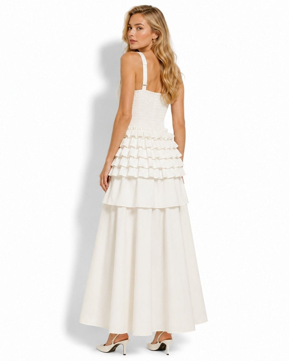 A woman modeling a long white tiered maxi dress from the back, featuring thin shoulder straps, a structured bodice, and a full flowing skirt, set against a plain white background.