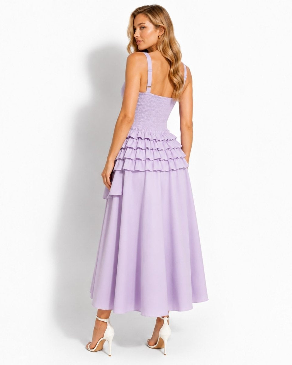 A woman modeling a lavender sleeveless maxi dress from the back, featuring a ruffled tiered waist and a flowing skirt, paired with white high heels against a white studio background.
