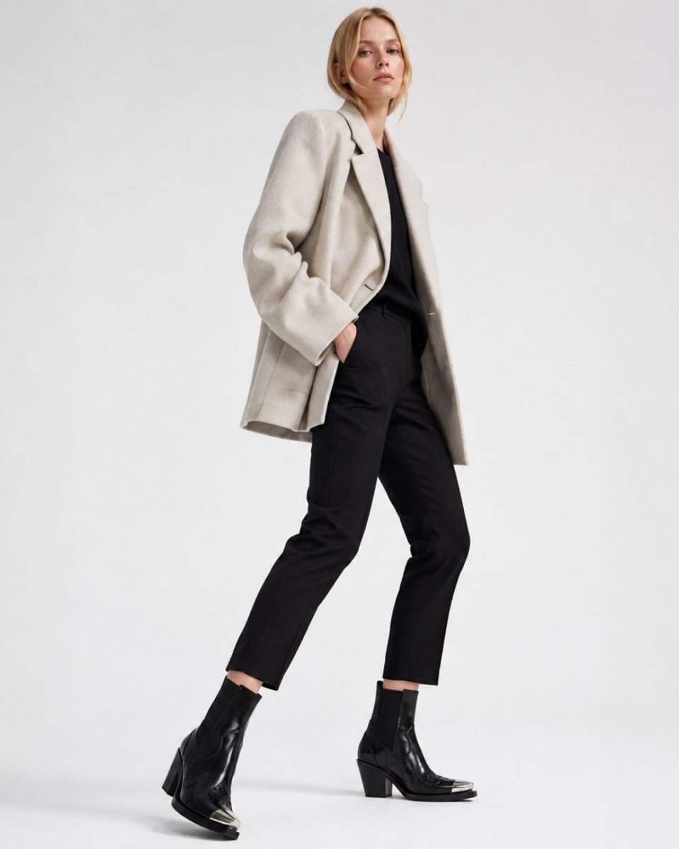 A model is shown from the waist down wearing a light beige oversized blazer, black trousers, a black top, and black leather ankle boots against a plain white studio background.