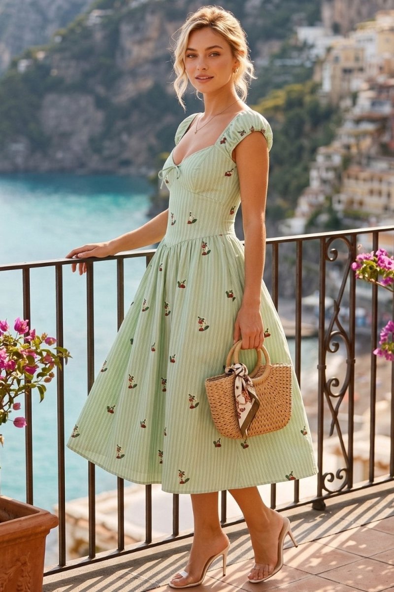 A woman stands on a balcony overlooking the blue sea and coastal town, wearing a light green pinstriped midi dress with a small floral print. She is holding a woven straw handbag with a pink ribbon and wearing nude high-heeled sandals.