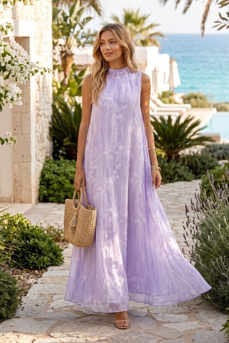 A woman stands on a stone walkway outdoors wearing a sleeveless lavender maxi dress with a high neck and flowing tulle skirt. She is holding a woven straw tote bag, with tropical plants, a pool, and blue ocean water in the background.