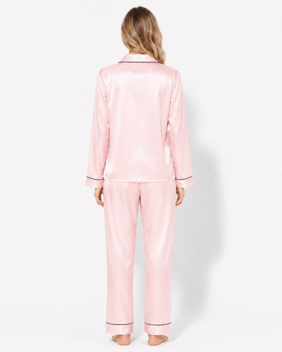 A woman viewed from behind wearing a light pink satin pajama set with thin dark piping trim on the collar, cuffs, and hems, against a plain white studio background.