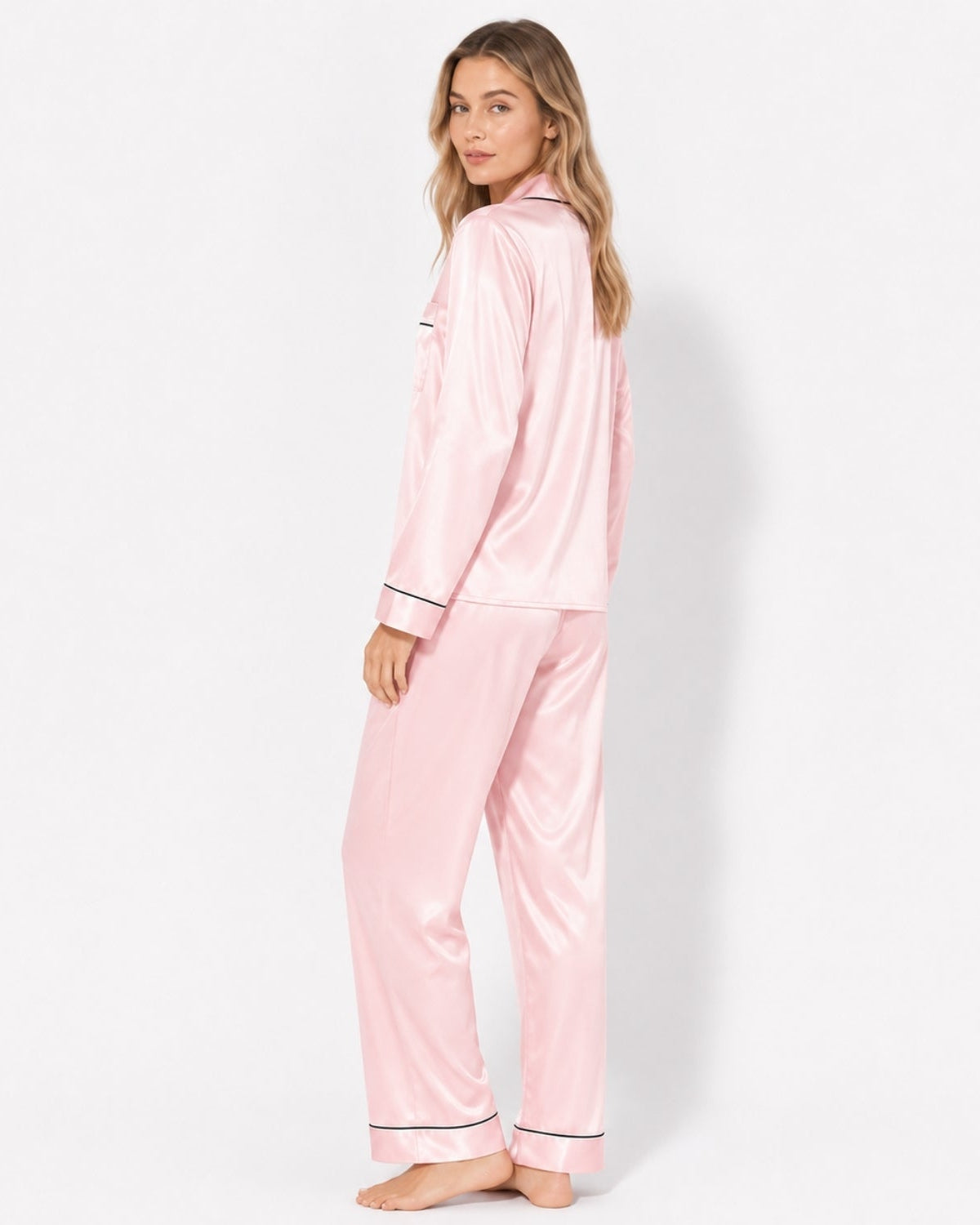 A woman modeling a light pink satin pajama set with black piping trim, shown from a side-back angle against a clean white studio background.