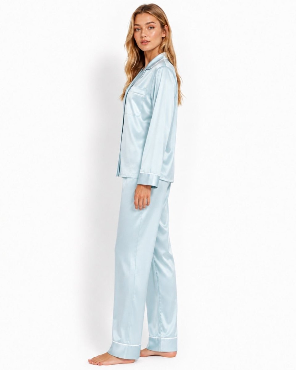 A woman modeling a light blue satin pajama set consisting of a long-sleeve button-up shirt and matching pants, standing against a plain white studio background.