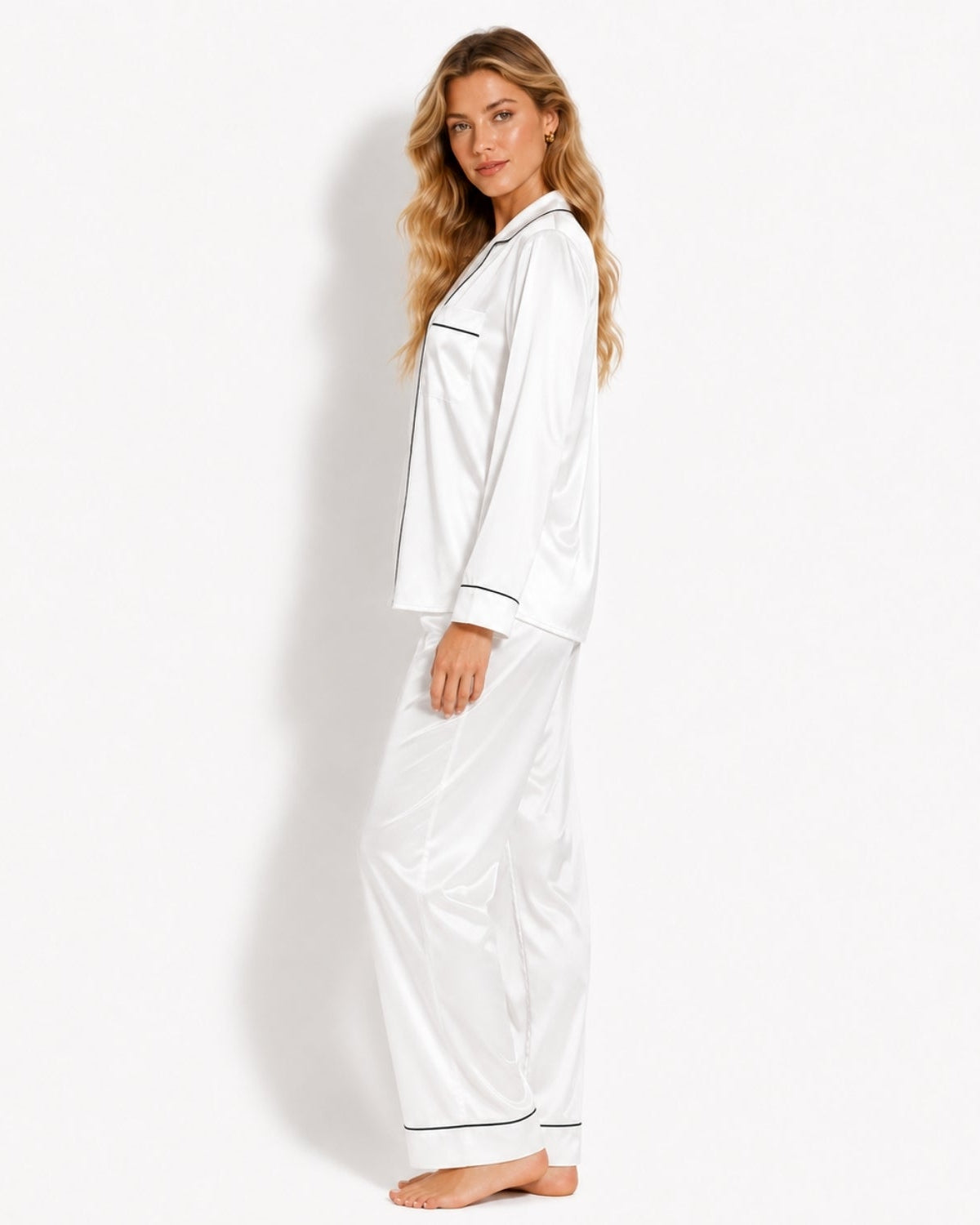 A woman modeling a white satin pajama set with black piping details, consisting of a long-sleeve top and matching pants, against a plain white studio background.