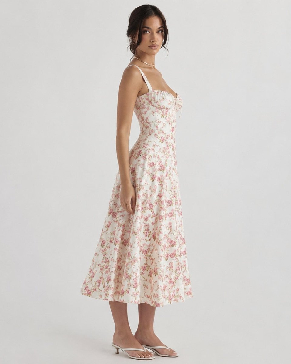 A woman modeling a sleeveless midi dress with thin straps and a pink floral print on a cream background, paired with clear high-heeled sandals, against a light gray studio background.
