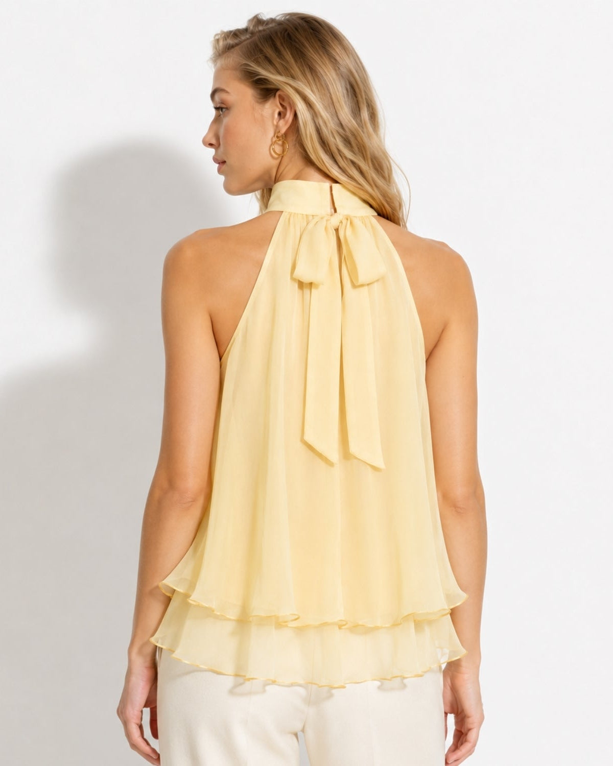 A woman shown from the back wearing a pale yellow halter top with a large bow at the neck and a layered ruffle hem, paired with light pants against a plain white studio background.