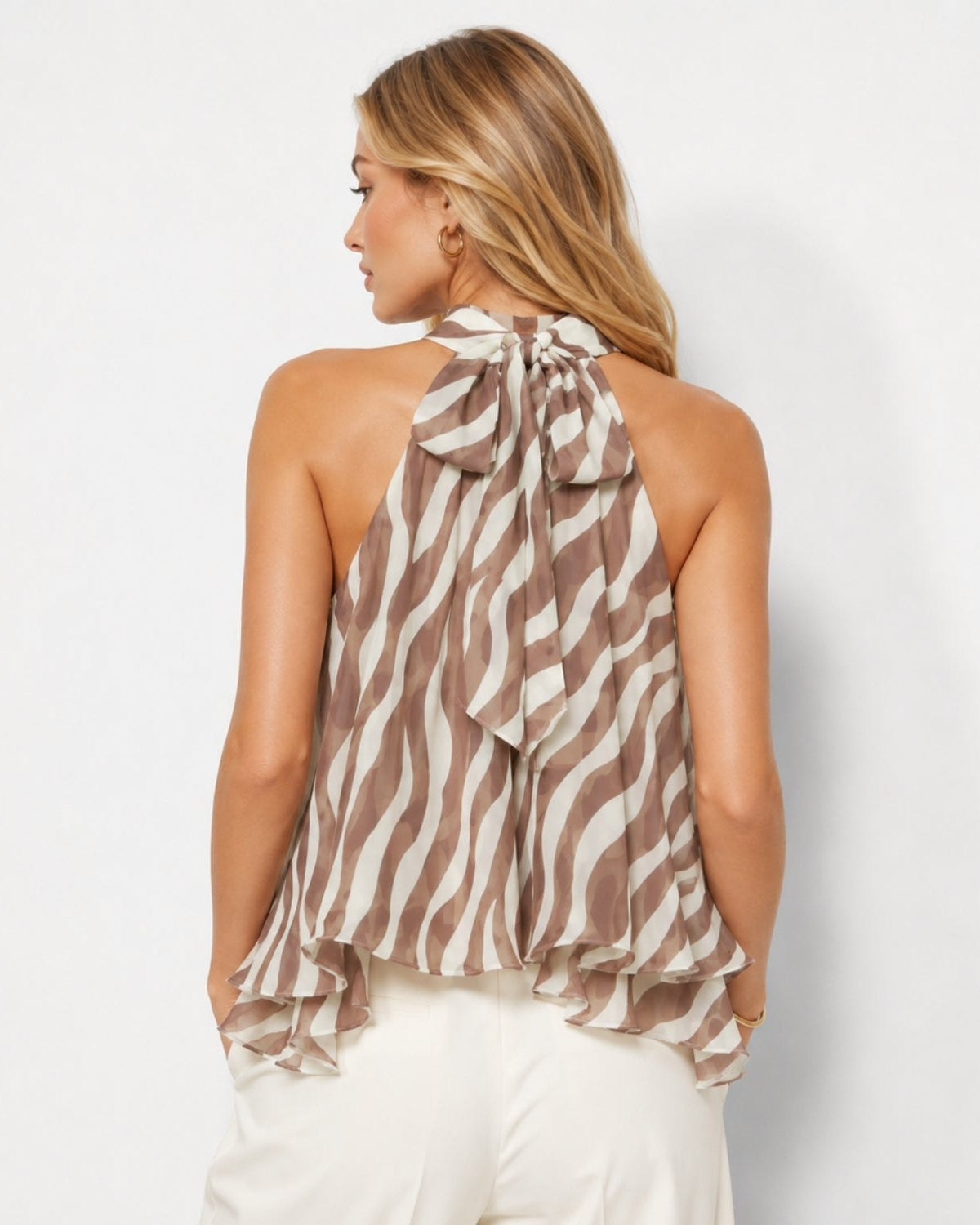 A woman shown from the back wearing a brown and cream zebra-print halter top with a large bow at the back, paired with white pants, against a light gray studio background.