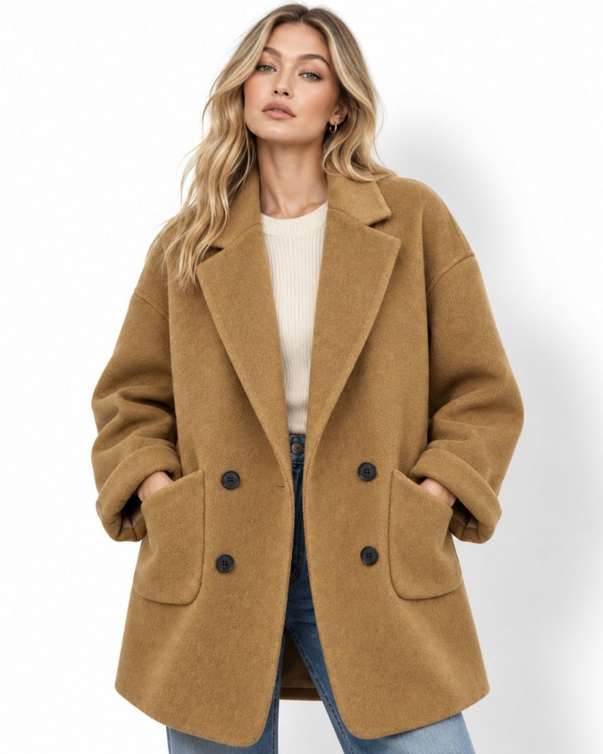 Woman wearing oversized double breasted amber peacoat over a white sweater and blue jeans, hands in patch pockets.