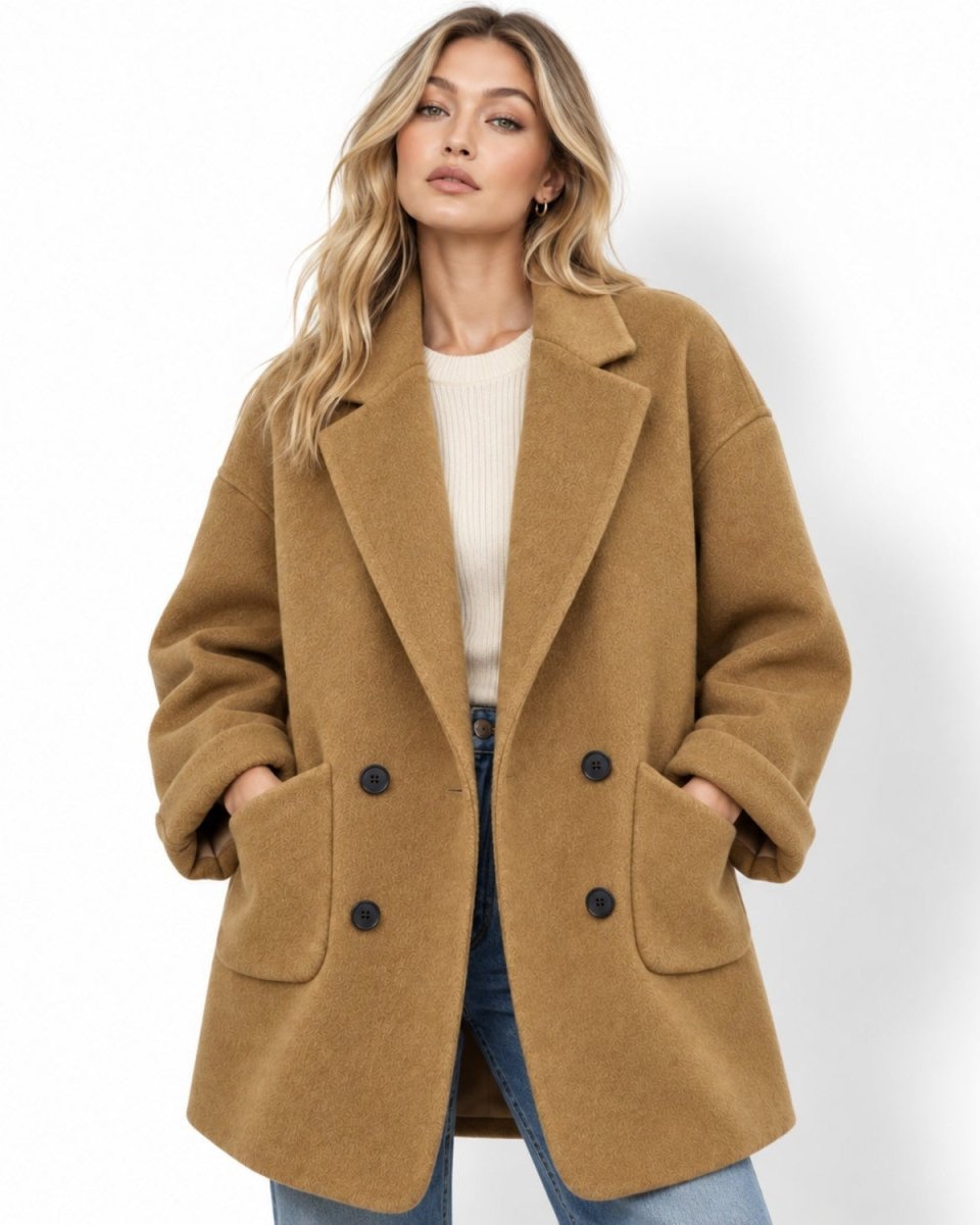 Woman wearing oversized double breasted amber peacoat over a white sweater and blue jeans, hands in patch pockets.