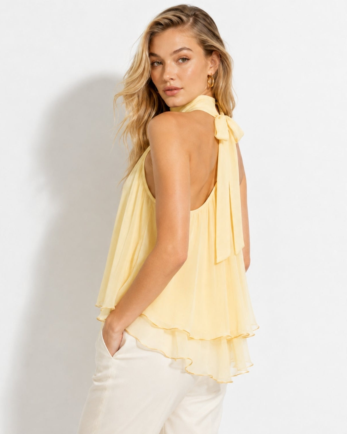 Side view of a woman modeling a arpicot sleeveless chiffon flowy top featuring an elegant back bow tie closure and layered hem.