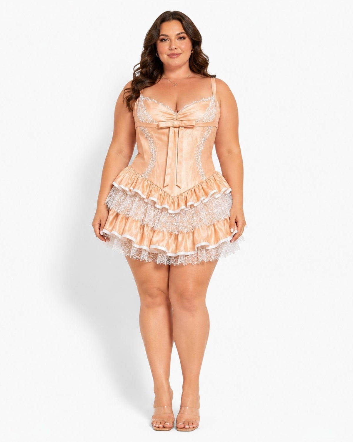 A woman modeling a peach satin corset-style mini dress with white lace trim and ruffles, standing against a plain white studio background.