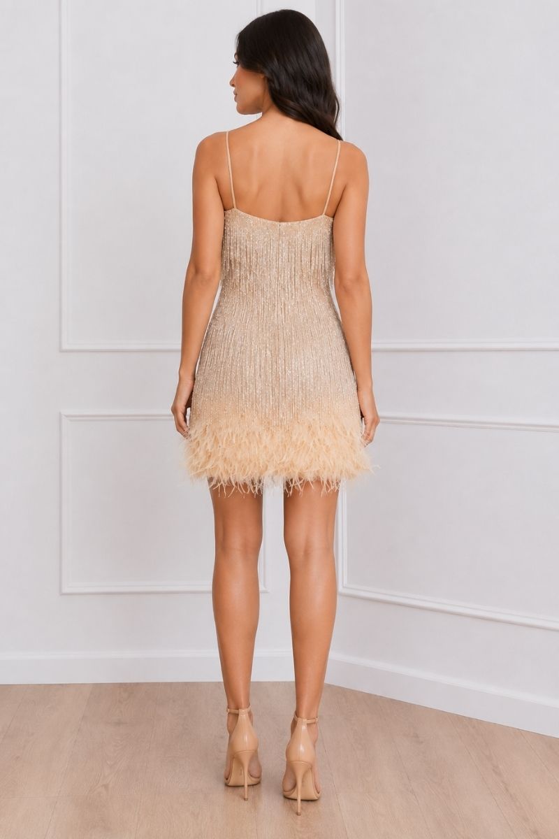 A woman viewed from the back wearing a sleeveless champagne-beige mini dress with thin straps, a textured beaded upper section, and a feathered hem, paired with nude high heels in a white paneled room.