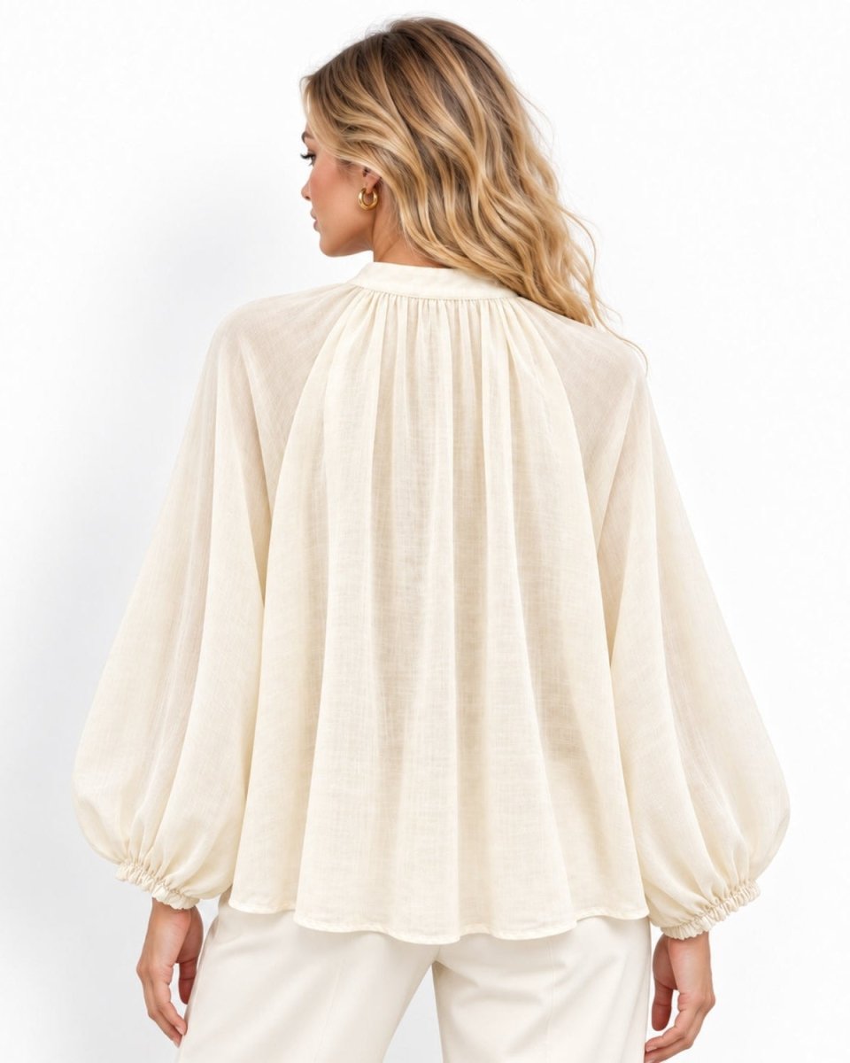 A woman shown from the back wearing a cream-colored long-sleeve blouse with a gathered, balloon-like silhouette and matching cream pants, set against a plain white studio background.