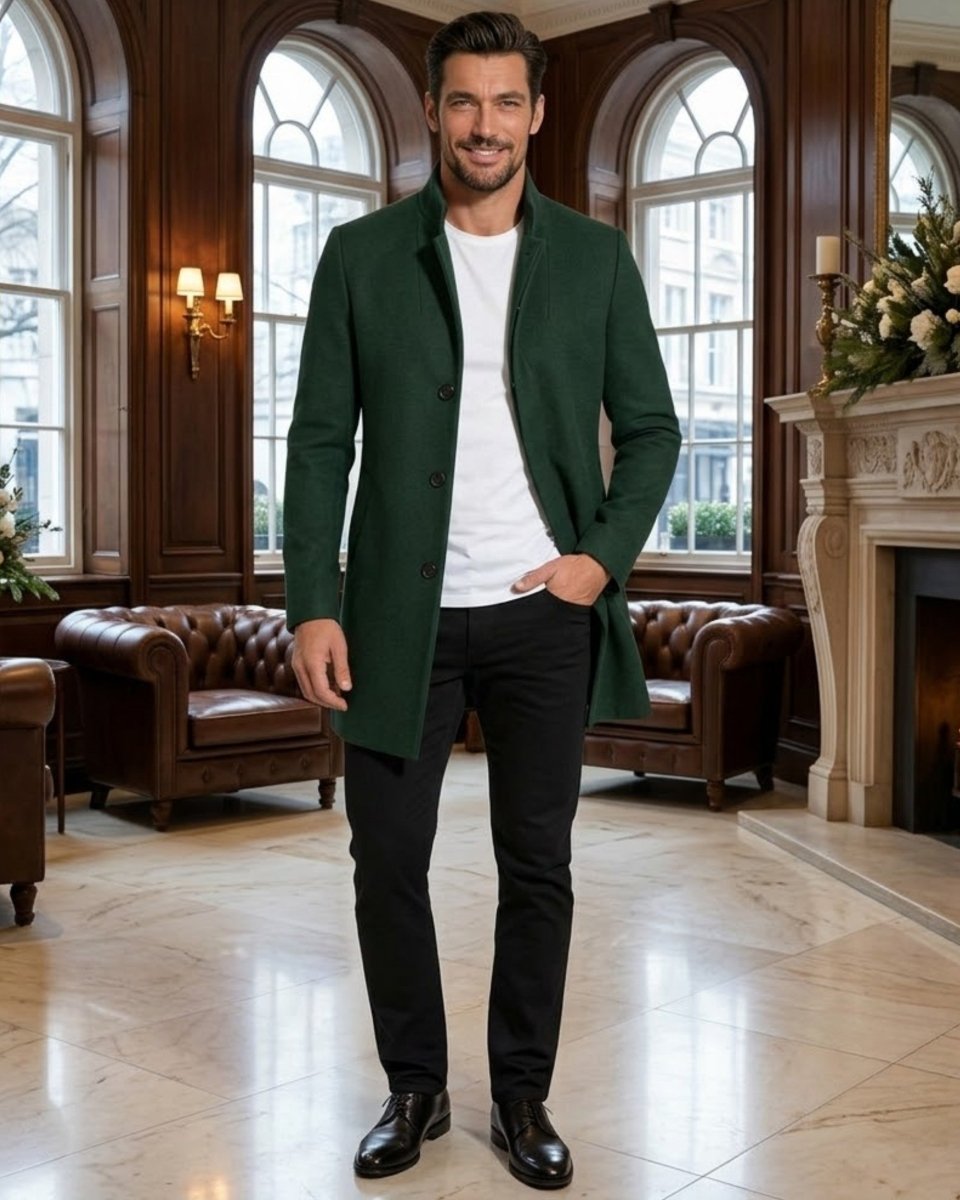 A man wearing a dark green wool-blend coat over a white t-shirt, black trousers, and black leather shoes, standing in a classic, elegant room with wood paneling, leather armchairs, and a fireplace.