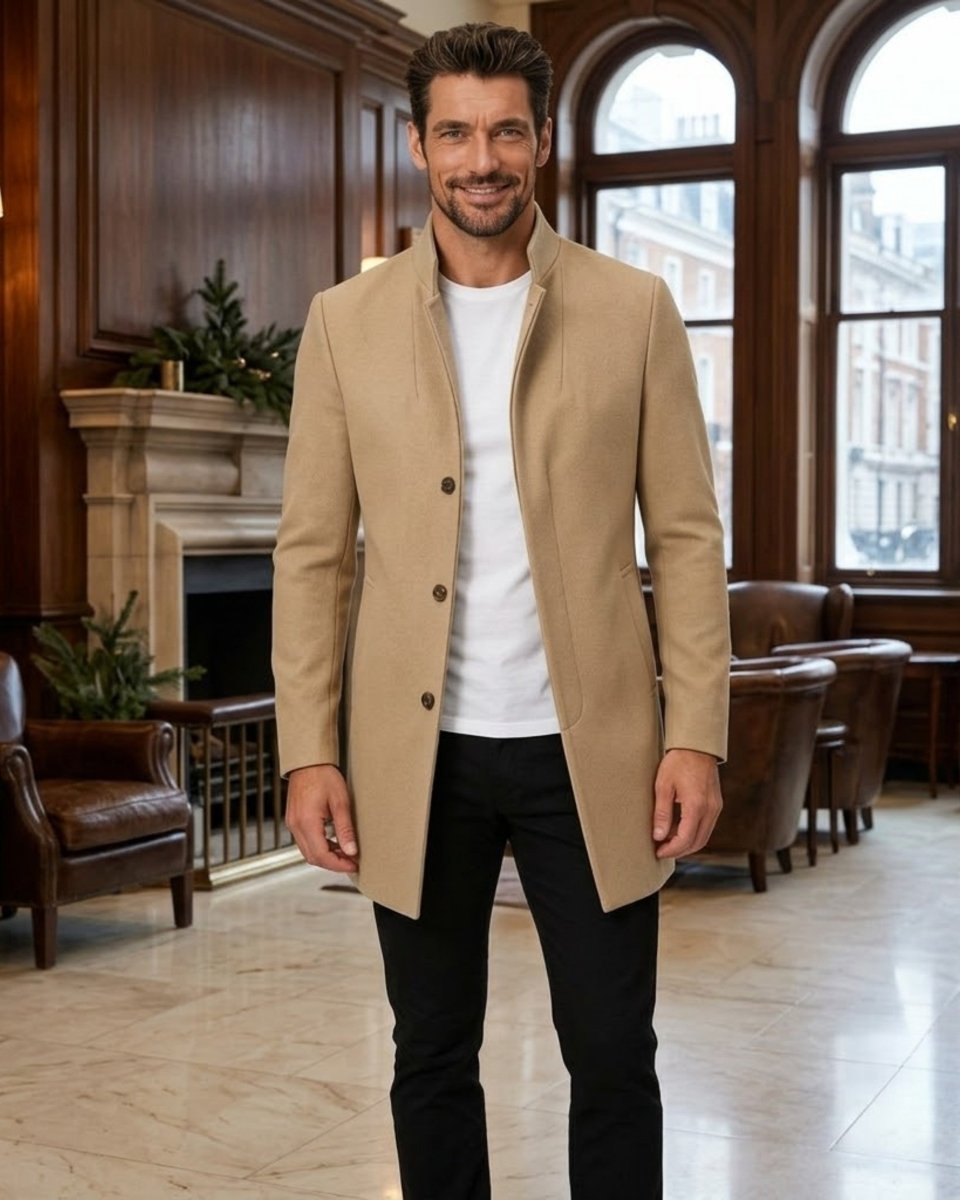A man wearing a beige mid-length blazer over a white t-shirt and black trousers, standing in a classic, elegant room with wood paneling, a fireplace, and large arched windows.