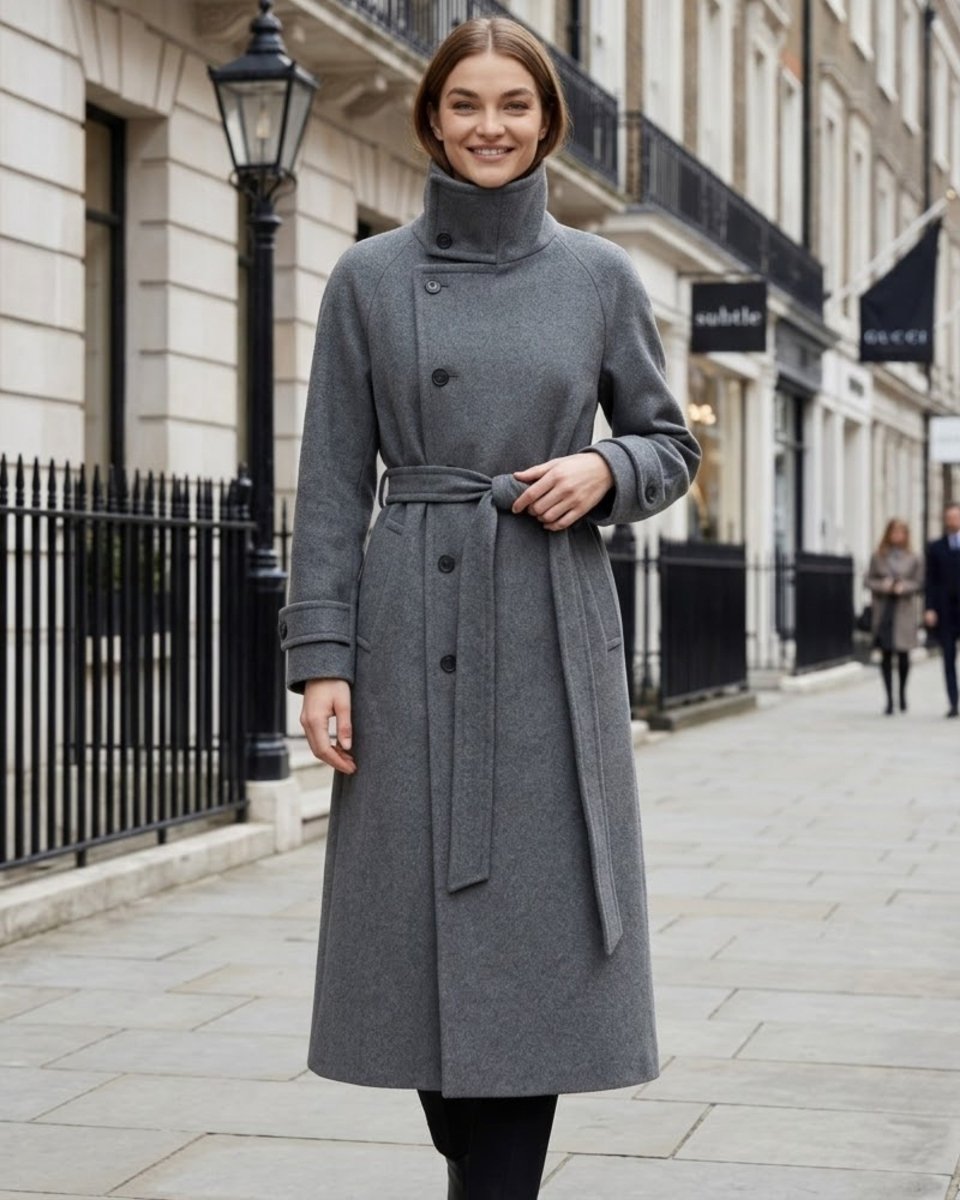 A woman wearing a mid-length gray wool coat with a high mock neck, double-breasted front, and matching waist tie belt, standing on a city sidewalk.