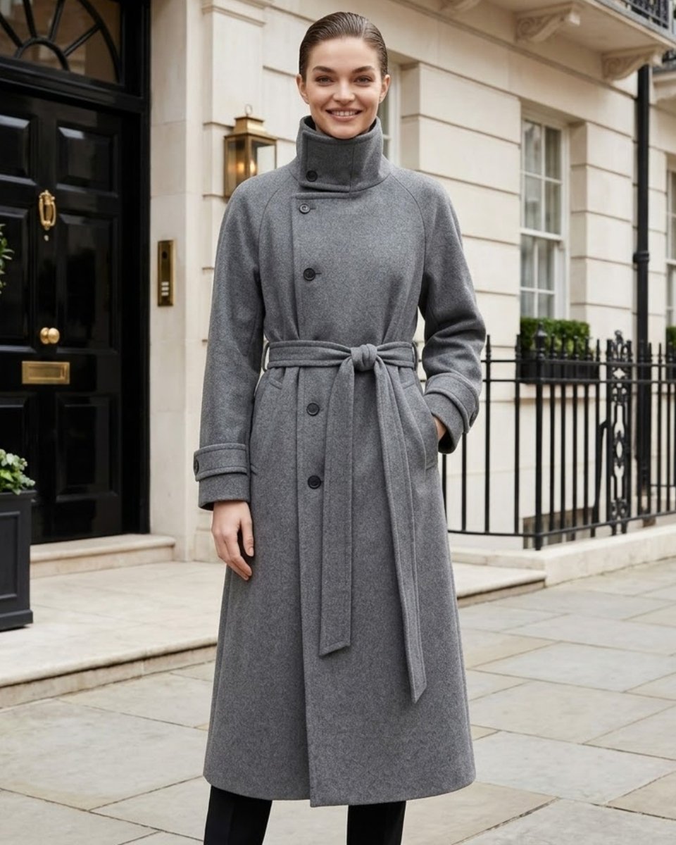 A woman wearing a mid-length gray wool coat with a high mock neck, double-breasted front, and matching waist tie belt, standing on a stone sidewalk in front of a classic cream-colored building with a black door.