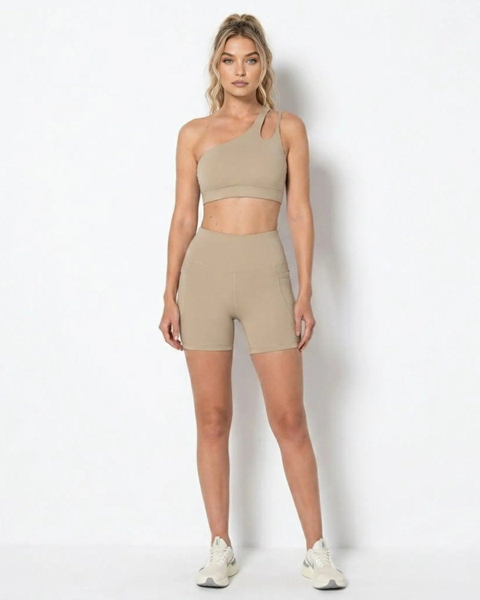 Blonde female model wearing khaki asymmetrical one shoulder sports bra and matching high waisted biker shorts with pockets facing forward.