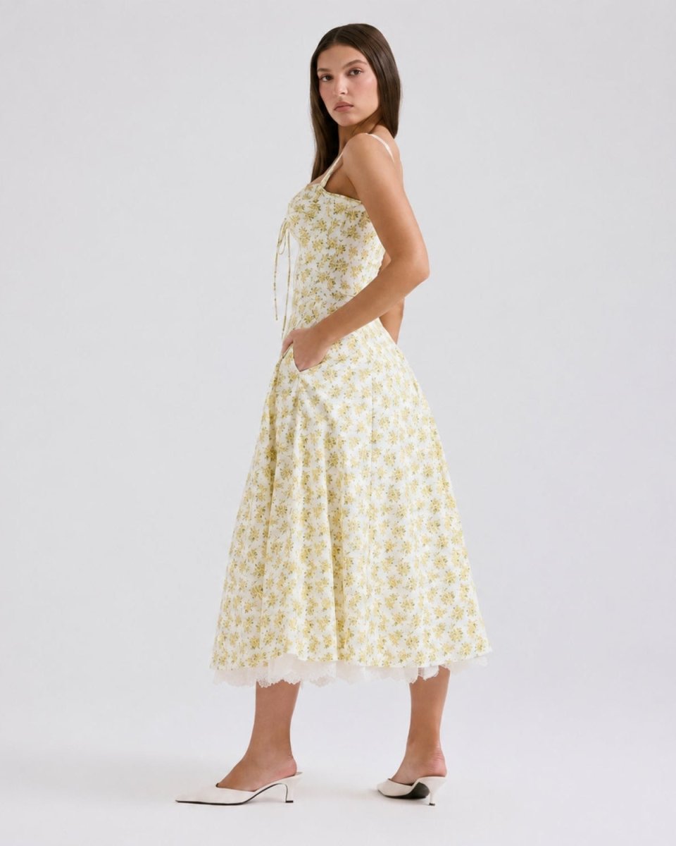 A woman modeling a sleeveless midi dress with a yellow and white floral pattern, standing against a light gray studio background.