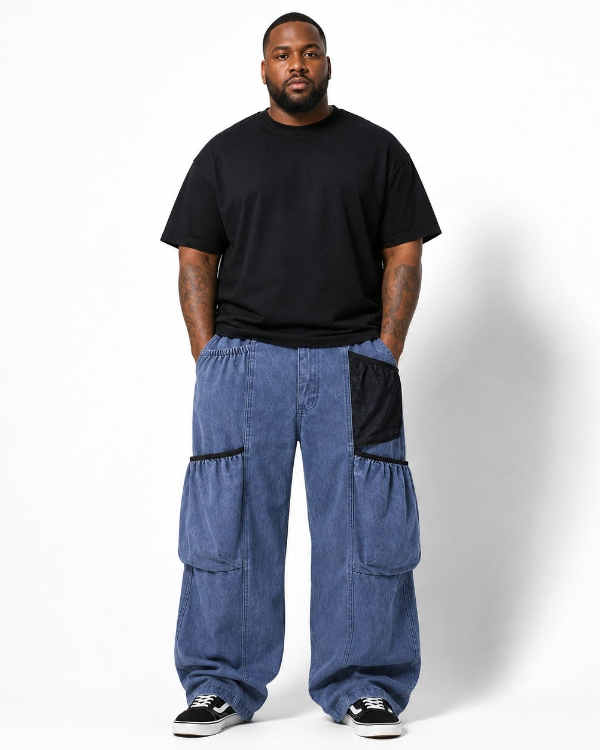 A man modeling a pair of baggy blue denim cargo pants with black contrast pocket details, paired with a black t-shirt and black sneakers, standing against a white studio background.