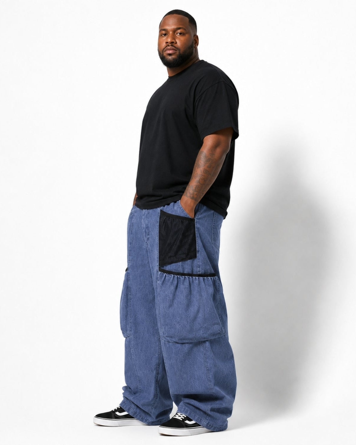 A man wearing a black t-shirt and baggy blue denim cargo pants with black mesh side pockets, standing against a white studio background.
