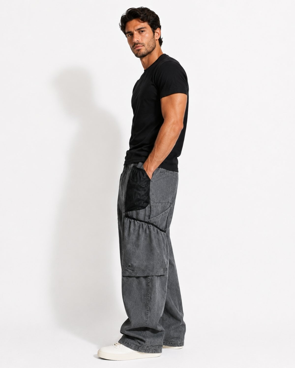 A man wearing a black t-shirt and dark gray wide-leg cargo pants with black patch pockets, standing in profile against a white studio background.