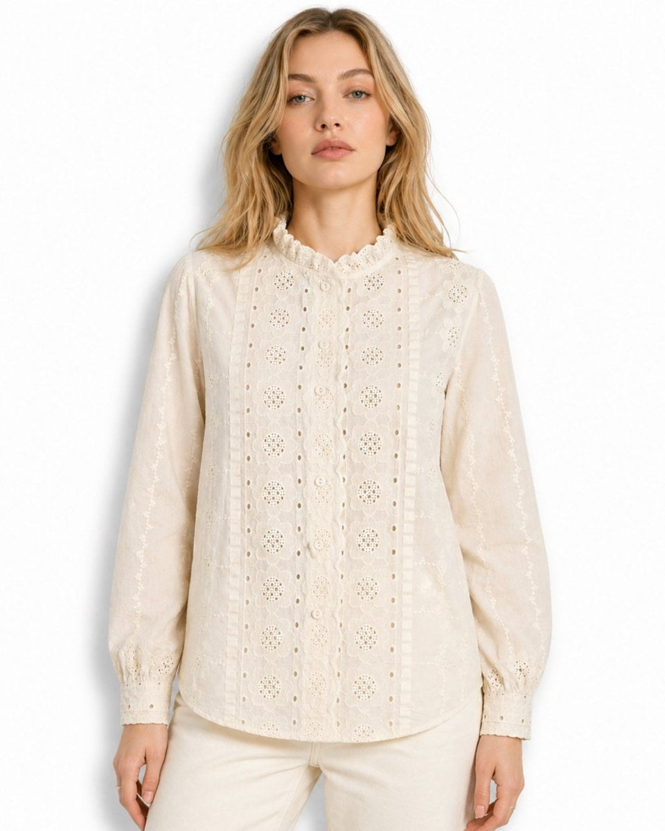 Front view of beautiful woman wearing an beige broderie anglaise long sleeve blouse with ruffled mock neck and floral eyelet embroidery against a white background.