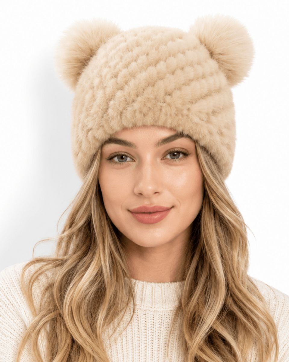 A woman wearing a beige chunky knit beanie with two fluffy pom-poms on top, paired with a cream knit sweater, against a plain white background.