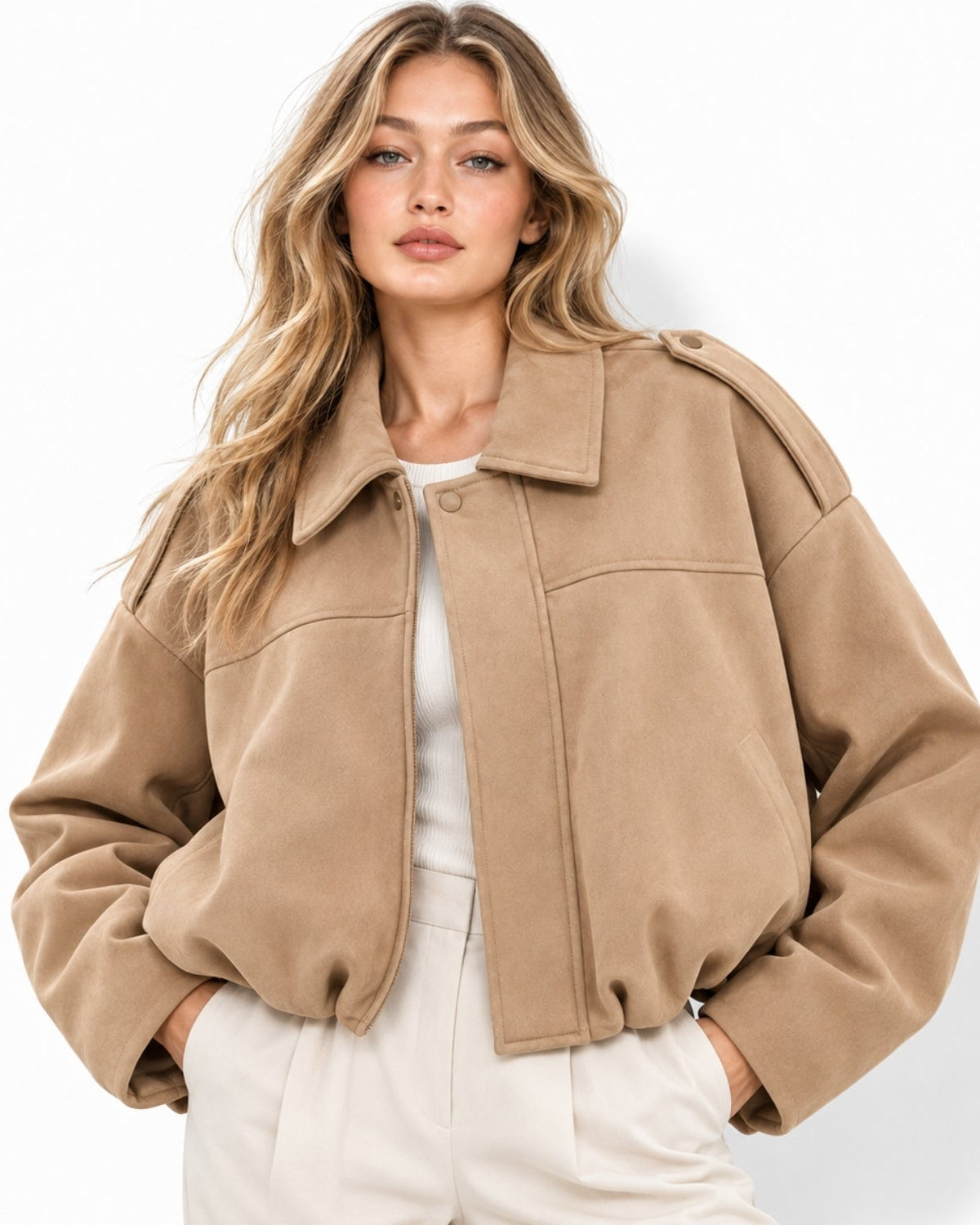 A woman wearing a cropped, oversized tan suede jacket over a white top and white pants, photographed against a clean white studio background.