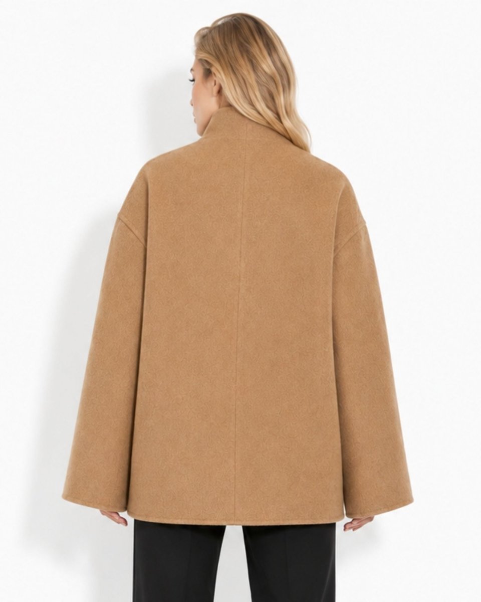 A woman viewed from the back wearing an oversized camel-colored coat with a high mock neck and wide sleeves, paired with black pants, against a plain white studio background.