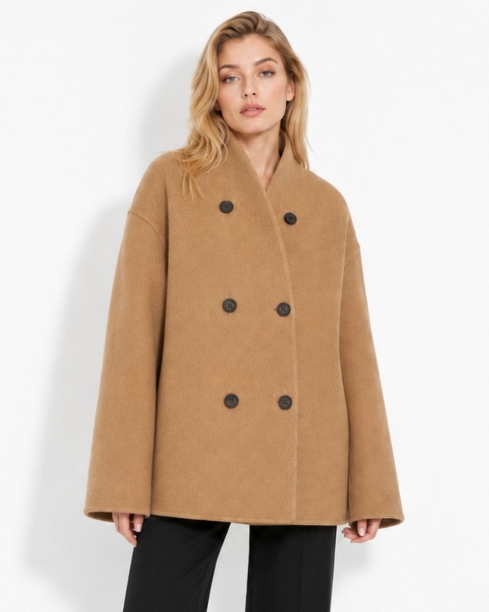 A woman modeling a camel-colored double-breasted coat with black buttons, paired with black pants, against a plain white studio background.