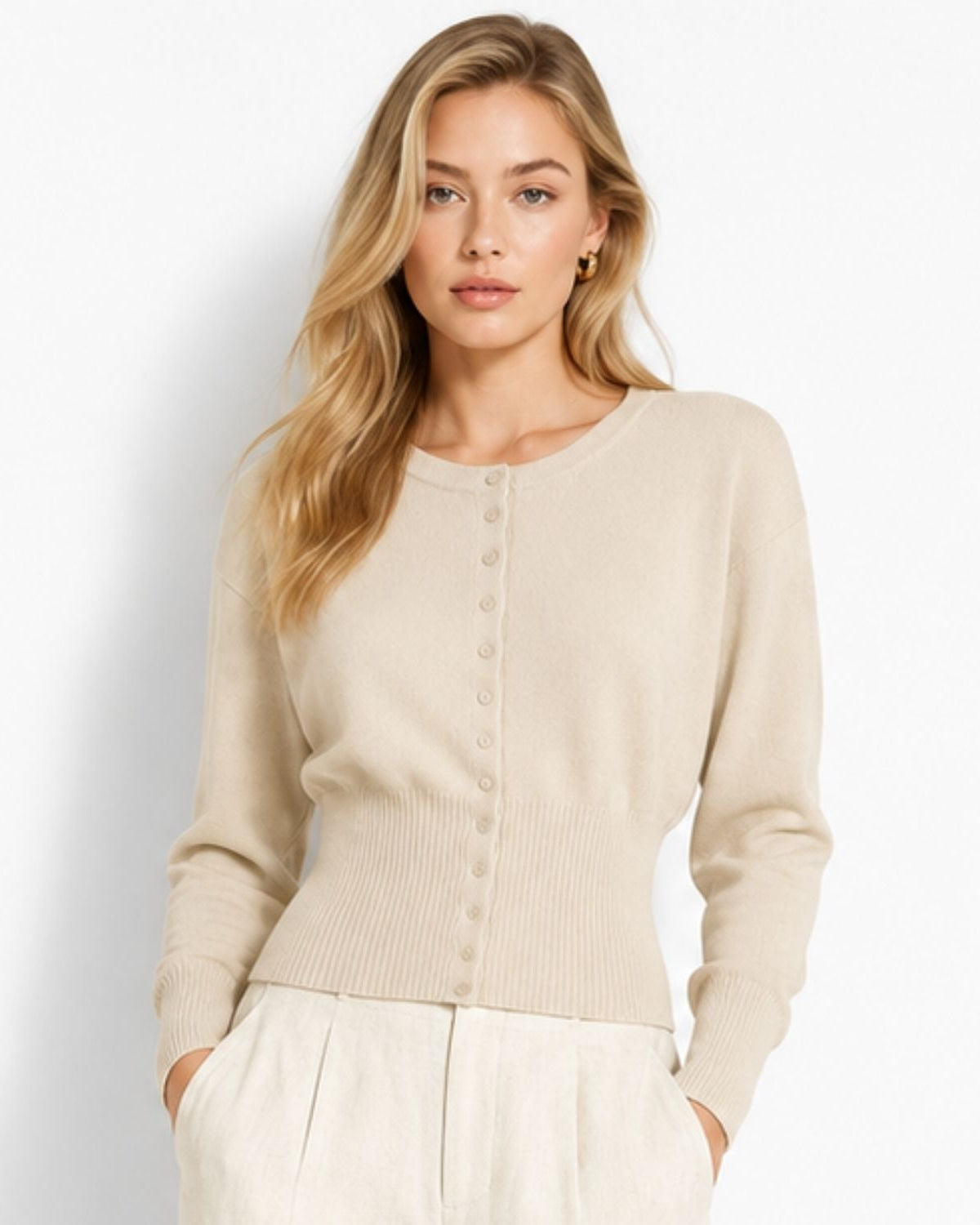 A woman modeling a beige crewneck knit cardigan sweater with a button-front closure, paired with off-white trousers, against a clean white studio background.