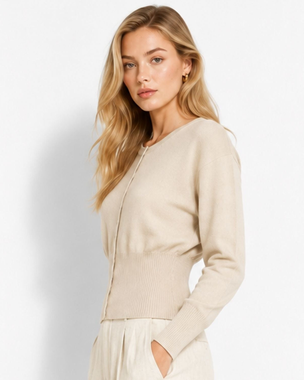 A blonde woman wearing a light beige knit cardigan sweater and matching cream trousers, standing against a clean white studio background.