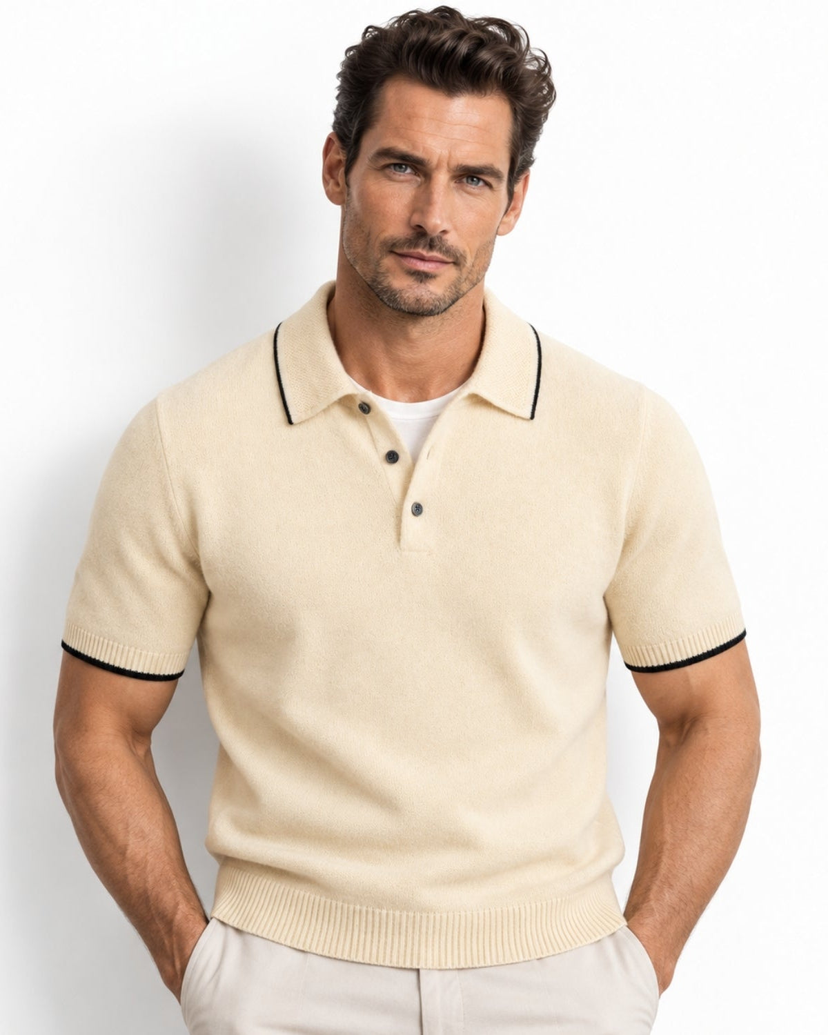 A man wearing a beige-colored short-sleeve knit polo shirt with black trim on the collar and sleeve cuffs, paired with light beige trousers, against a plain white studio background.