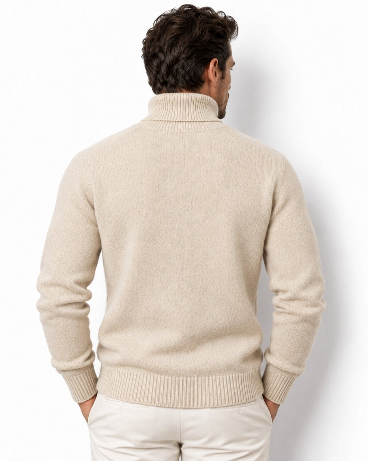 A man viewed from behind wearing a light beige knit turtleneck sweater and off-white trousers, standing against a plain white studio background.