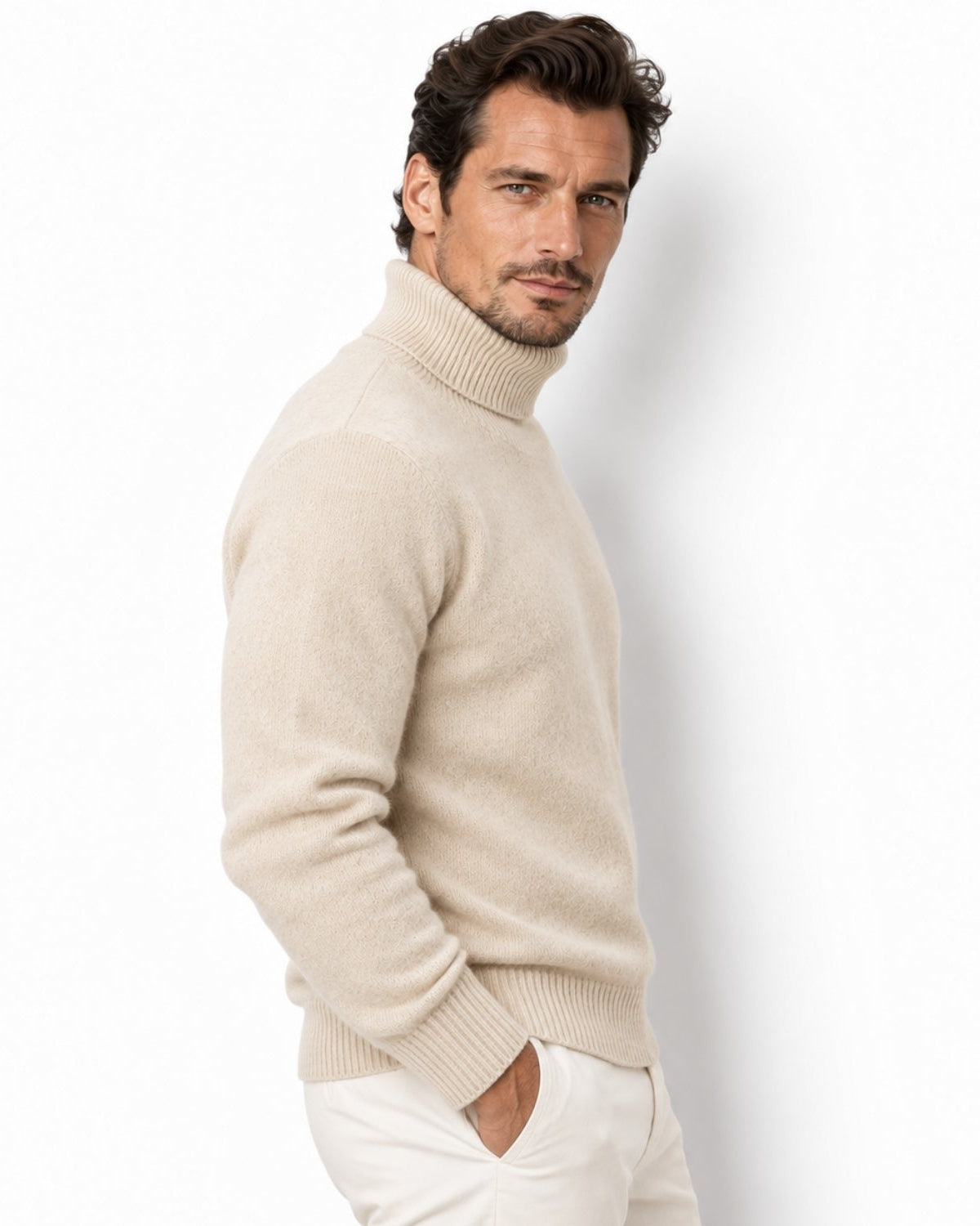 A man wearing a light beige knit turtleneck sweater and white trousers, standing against a plain white studio background.