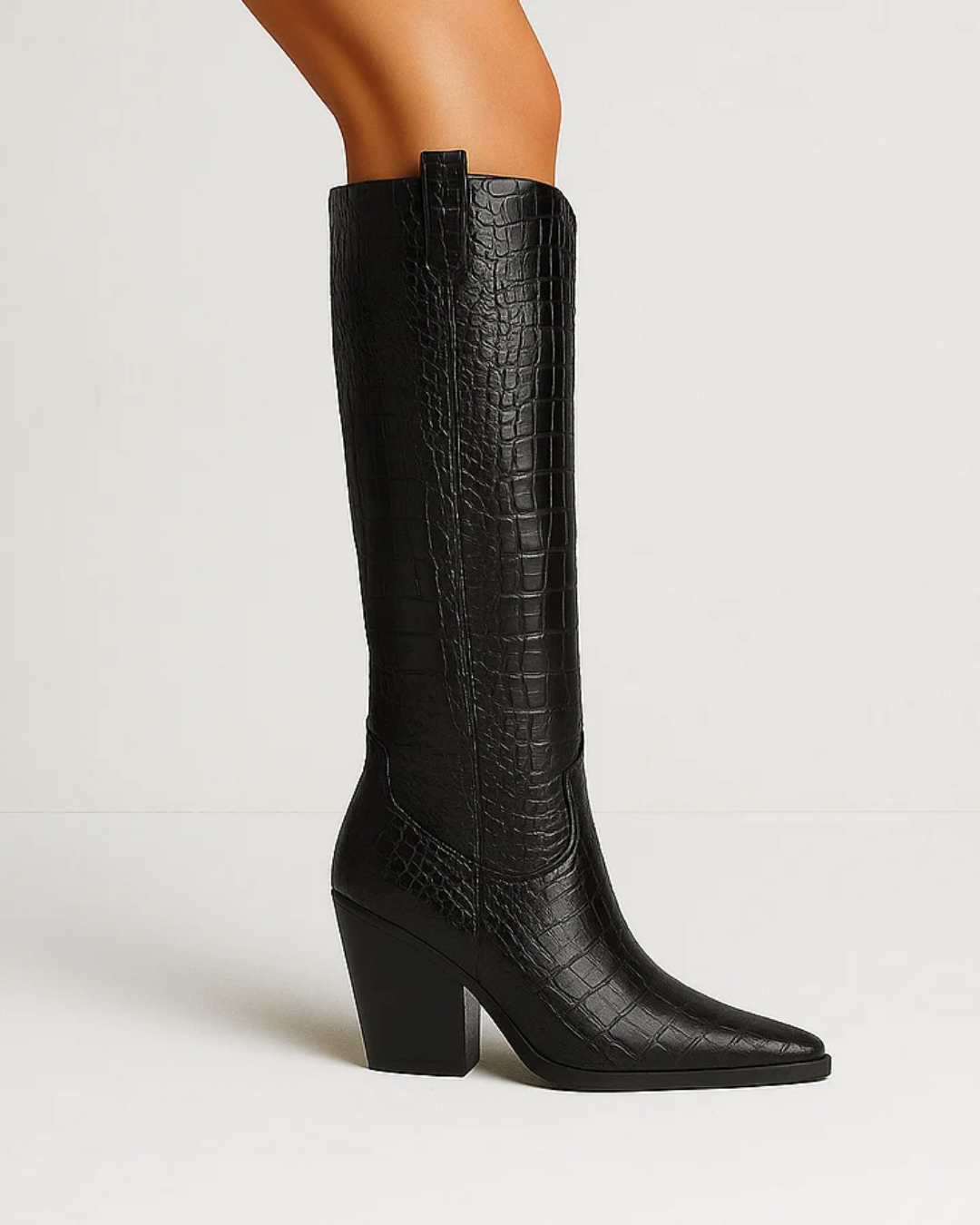 A single black knee-high boot with a crocodile-embossed texture, featuring a pointed toe and a chunky block heel, shown on a model's leg against a plain white studio background.