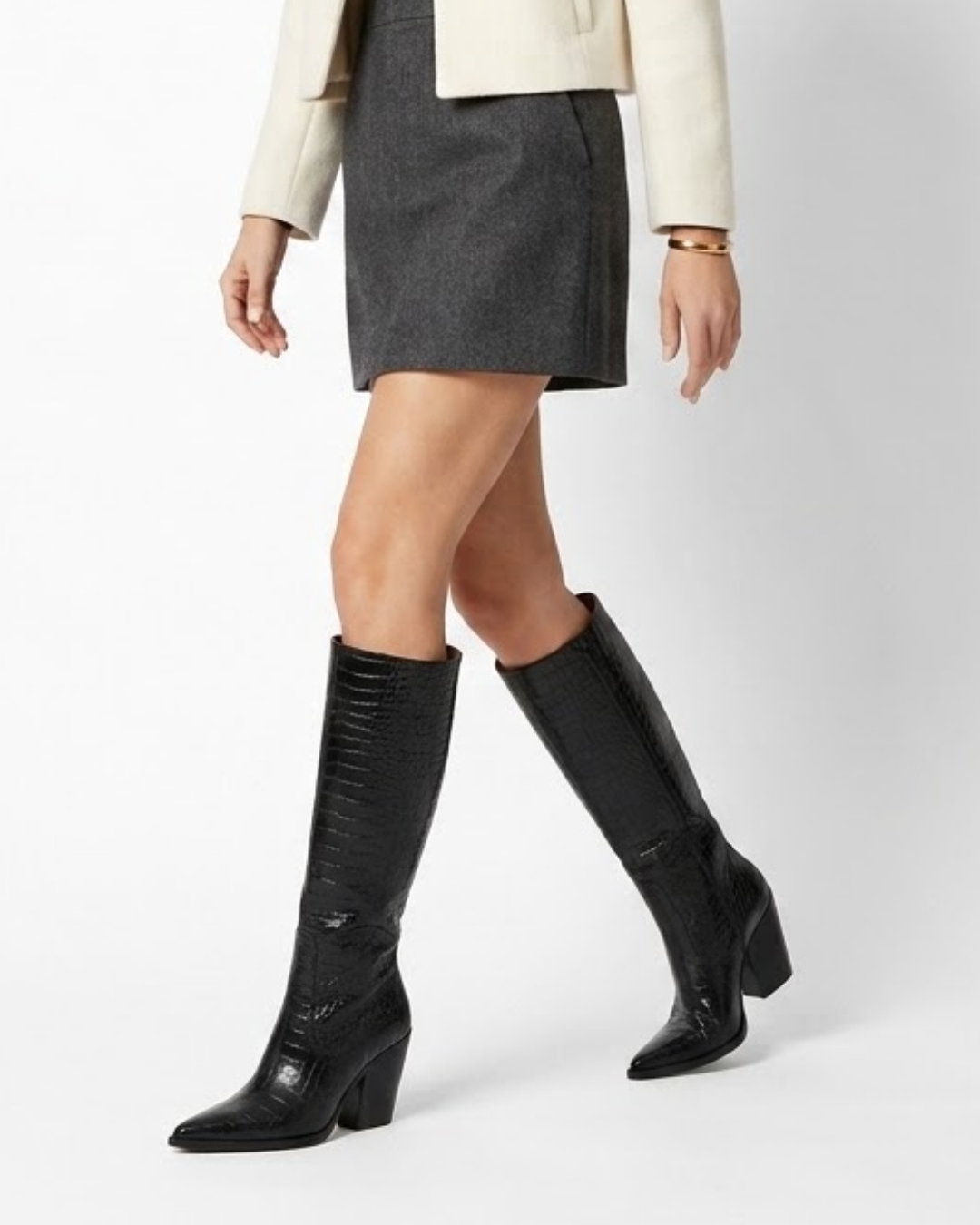 A model wearing a pair of black knee-high boots with a textured crocodile-embossed finish, paired with a dark gray mini skirt and a cream blazer, shown from the waist down against a light gray studio background.