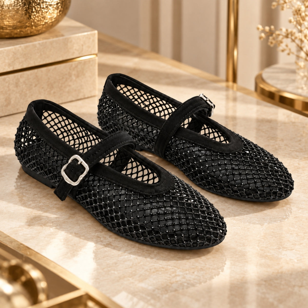 A pair of black mesh Mary Jane flats with black straps and silver buckle details, displayed on a beige marble surface with gold-toned decorative props in the background.