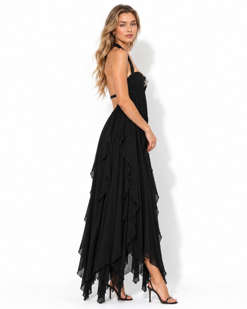 A woman modeling a black halter-neck maxi dress with a ruffled, tiered skirt and asymmetrical hemline, paired with black high-heeled sandals against a plain white studio background.