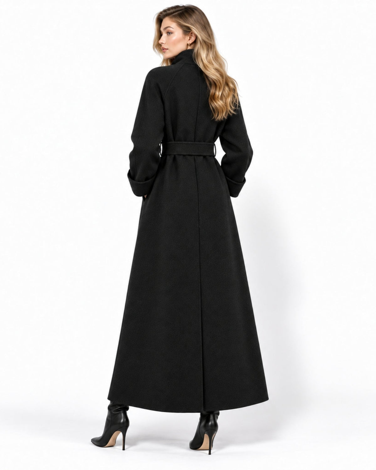 A woman modeling a long black coat from the back, featuring a high collar, long sleeves, and a matching waist belt, paired with black high-heeled boots against a plain white studio background.