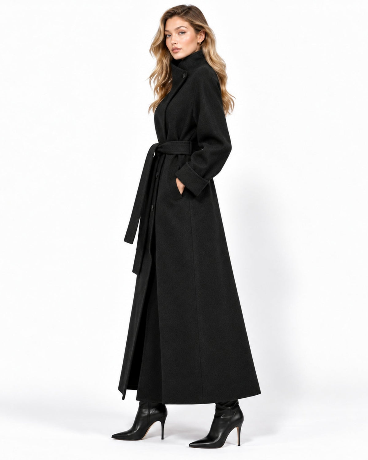 A woman modeling a long black wrap-style coat with a waist tie and wide lapels, paired with black ankle boots, against a plain white studio background.