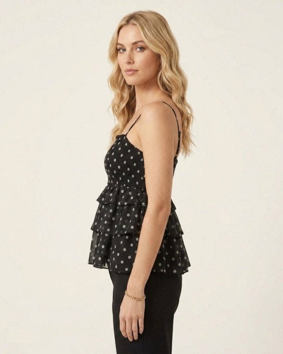 A blonde woman wearing a black spaghetti-strap peplum top with a white polka-dot pattern, paired with black pants, photographed against a plain off-white studio background.