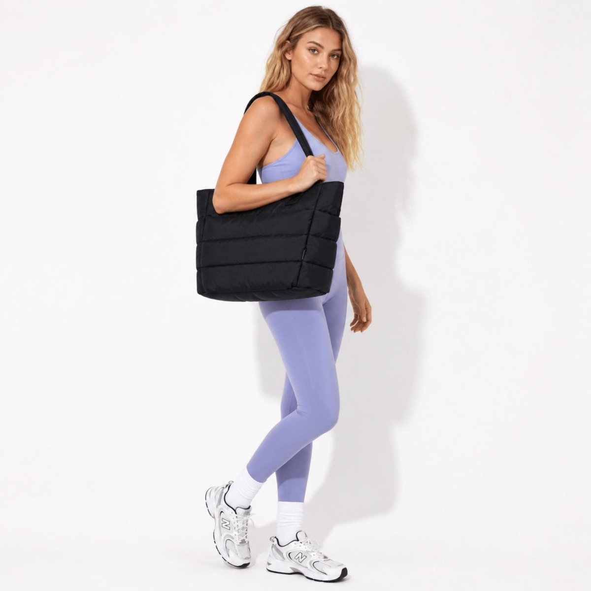 A woman in a lavender athletic set carrying a large black puffer tote bag against a clean white studio background.
