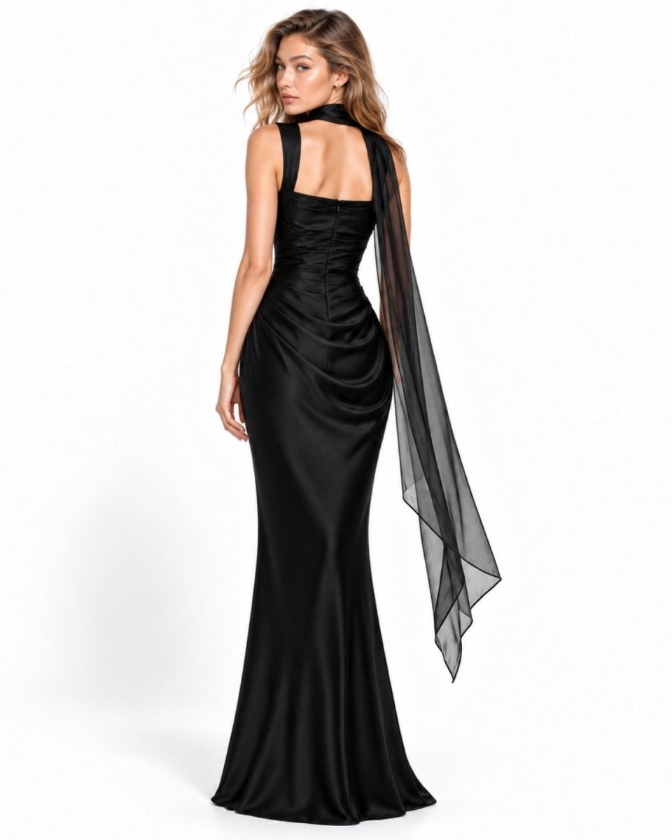 A woman modeling a floor-length black satin gown with a draped sheer black overlay, shown from the back against a clean white studio background.