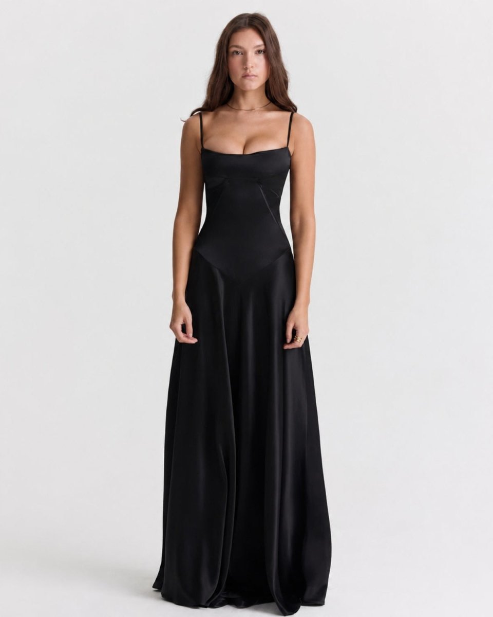 A woman modeling a floor-length black satin slip dress with thin spaghetti straps and a sweetheart neckline, photographed against a seamless light gray studio background.