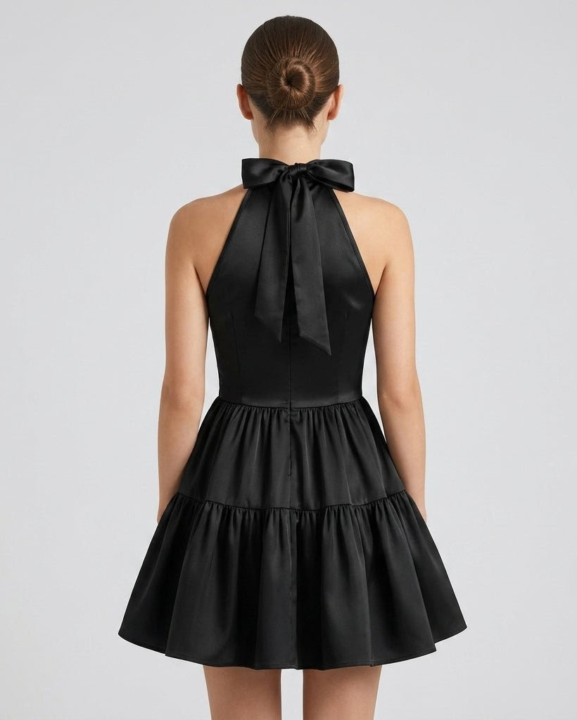 Back view of model wearing the black satin little black dress, showcasing the open shoulder design, back zipper, and elegant neck tie bow.