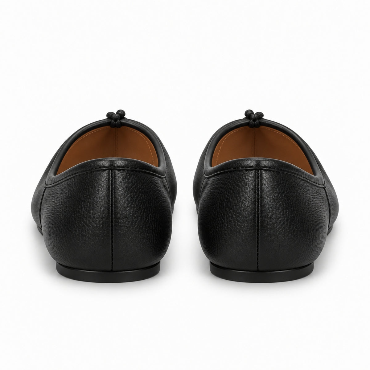 A pair of black pebbled leather ballet flats shown from the back, featuring a small black tassel detail on the back of the shoe and a contrasting tan interior lining, set against a plain white background.